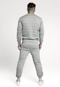 Grå mönstrad sweatshirt och joggers-set med ribbad fåll; mjuk textur; elastiska muddar; vita sneakers synliga vid fötterna.