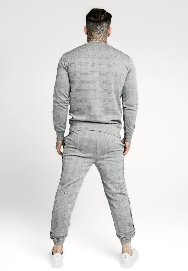 Grå mönstrad sweatshirt och joggers-set med ribbad fåll; mjuk textur; elastiska muddar; vita sneakers synliga vid fötterna.