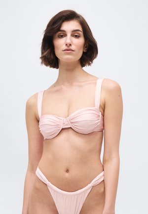 Νέα γυναίκα με κοντά καστανά μαλλιά, φορώντας ροζ ανοιχτό ρουχαριστό τοπ μπικίνι και αντίστοιχα μαγιό με ψηλή μέση, σε απλό φόντο.