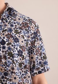 Camicia a maniche corte con un motivo floreale in navy, marrone e blu su uno sfondo bianco. Realizzata in tessuto leggero con un design colletto.