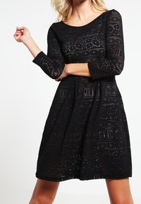 Femme portant une robe noire longueur genoux à manches longues avec un motif découpé géométrique ressemblant à de la dentelle et un col rond, se tenant devant un fond blanc.