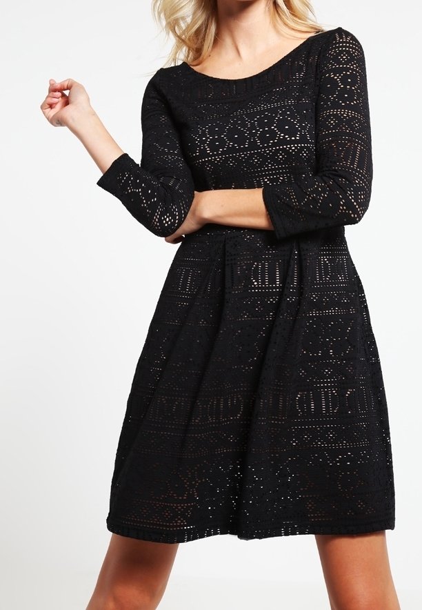 Femme portant une robe noire longueur genoux à manches longues avec un motif découpé géométrique ressemblant à de la dentelle et un col rond, se tenant devant un fond blanc.