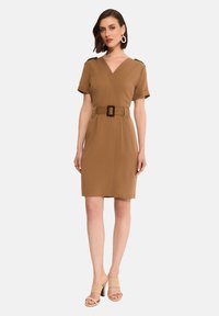 Brązowa sukienka z paskiem, z dekoltem w kształcie litery V, krótkimi rękawami i akcentami na ramionach. Gładki materiał z dopasowanym krojem. Noszona z beżowymi sandałami na wysokim obcasie.