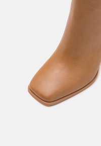 Gros plan sur une botte en cuir beige avec un bout carré et une texture lisse sur un fond blanc.