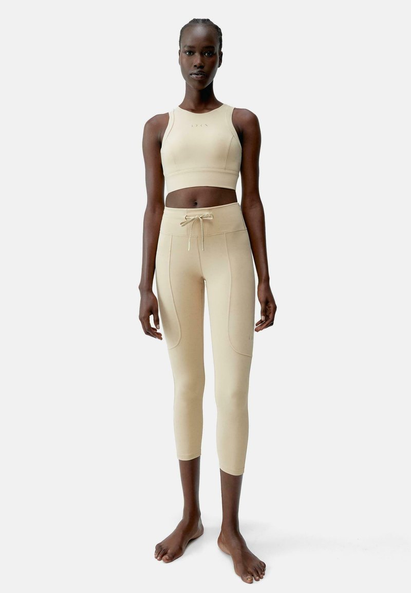 Bež športni crop top in legice iz raztegljivega materiala, ki imata prilegajoč dizajn, pas na vrvico in subtilne detajle šivov.