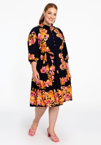 Blumenmusterkleid mit marineblauem Grund, lebhaften rosa und orangenen Mustern, gerafftem Ausschnitt, Dreiviertelärmeln und Taillenknotengürtel. Rosa Blockabsatzsandalen.