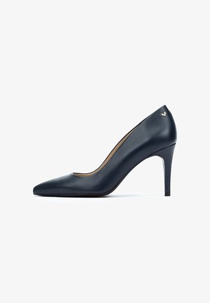 Zapato de tacón alto de cuero azul marino con puntera puntiaguda, tacón de aguja delgado y sutil acento de logo dorado en la parte posterior.
