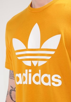 Personne portant un t-shirt orange avec un grand logo blanc Adidas trefoil et un texte sur la poitrine, montrant partiellement un bras tatoué.