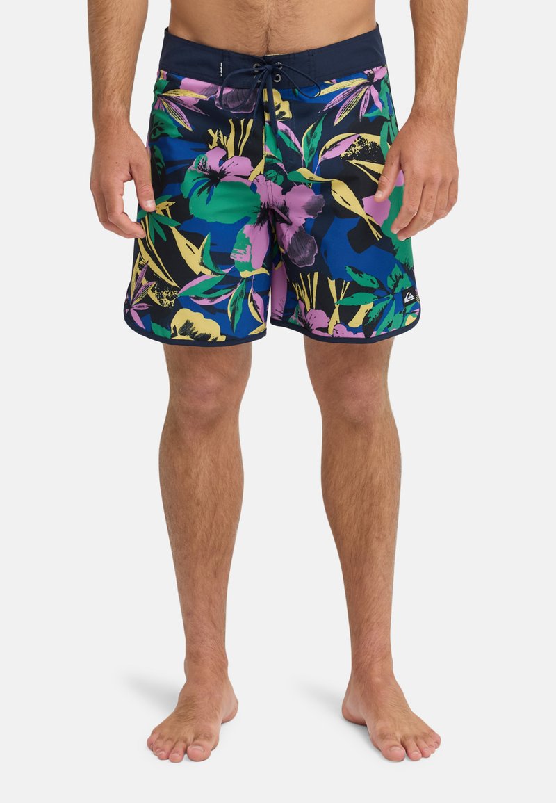 Mann trägt lebhafte, geblümte Badeshorts mit Kordelzug am Bund und steht barfuß vor einem schlichten weißen Hintergrund.