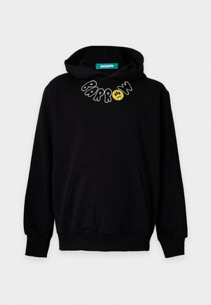 Homme portant un sweat à capuche noir avec le texte "Parron" et un visage souriant, jean bleu ample et baskets noires, debout les mains dans la poche du sweat.