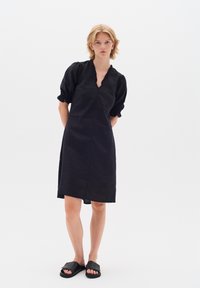 Schwarzes Leinenkleid mit V-Ausschnitt, puffigen Ärmeln und lockerer Passform. Der Saum ist hinten etwas länger und wird mit flachen schwarzen Sandalen kombiniert.