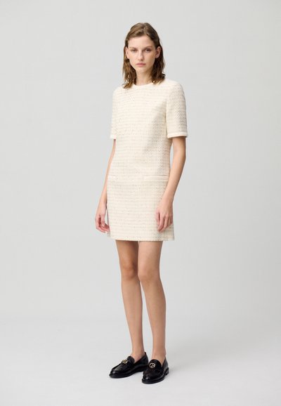 Robe crème à manches courtes avec motif texturé et poches latérales, fabriquée en tissu tissé. Mocassins noirs avec accents en métal doré.