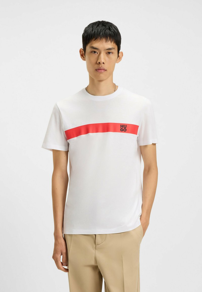 T-shirt in cotone bianco con una striscia orizzontale rossa sul petto e il logo "HUGO" in nero. Maniche corte, design a collo rotondo.