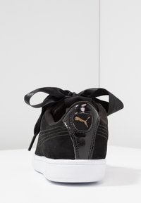 Baskets Puma en daim noir avec panneau arrière brillant, logo doré, grands lacets en satin noir noués en nœud, et semelle blanche texturée sur une surface blanche.