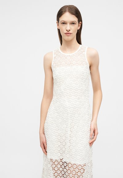 Jeune femme aux cheveux bruns lisses portant une robe blanche sans manches en dentelle avec un motif floral, debout devant un fond clair uni.