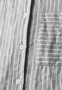 Camicia di lino grigia con strisce verticali bianche, chiusura con bottoni e una singola tasca applicata con motivo a strisce.