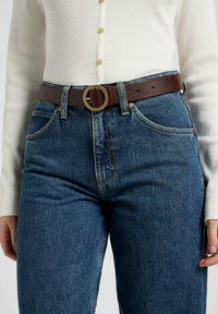 Cinturón de cuero marrón con una hebilla dorada redonda, llevado sobre unos pantalones vaqueros de tiro alto azules. Parte superior de manga larga blanca con relieve visible por encima.