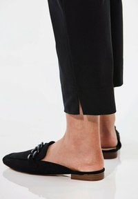 Pantalons noirs cropped avec des fentes sur les côtés à l'ourlet, associés à des chaussures noires à enfiler avec un détail de chaîne décorative et une semelle en bois plate.