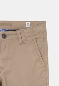 Pantalon beige pour enfants avec passants de ceinture, fermeture à bouton, poche latérale et étiquette de taille 152/12 ans à l'intérieur de la taille.