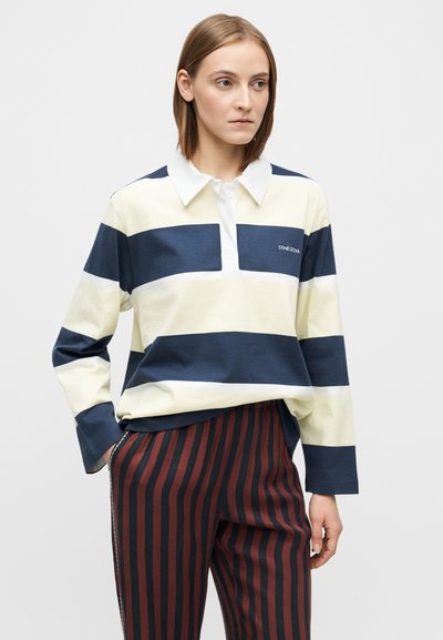 Tricou polo cu mâneci lungi, în dungi bleumarin și crem, având un guler alb și un detaliu cu logo pe partea stângă. Asortat cu pantaloni în dungi roșii și nuanțe închise.