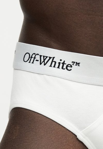 Gros plan sur une peau foncée portant un slip blanc avec une large ceinture présentant le logo noir "Off-White™".