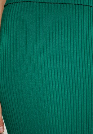 Grüner gerippter Stoff mit vertikalen Linien, der eine weiche Textur und ein tailliertes Design aufweist. Eine Nahaufnahme hebt die Textur und Farbe hervor.