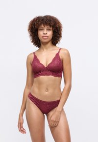 Donna con capelli ricci che indossa un reggiseno e una mutanda in pizzo bordeaux abbinati, in piedi contro uno sfondo semplice e chiaro.
