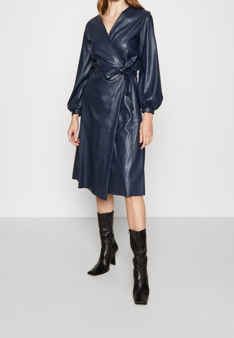 Femme portant une robe portefeuille en similicuir bleu marine avec une taille nouée et des manches longues bouffantes, associée à des bottes noires à talons mi-mollet.