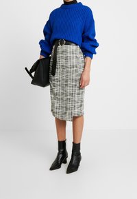 Blå ribbad tröja, grå rutiga pennkjol med svarta accenter, svart bälte och spetsiga svarta ankelskor. Svart väska hålls i vänster hand.