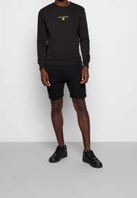 Svart sweatshirt med rund hals och långa ärmar, prydd med en gul "New Horizon"-logotyp. Matchas med svarta shorts och svarta sneakers.