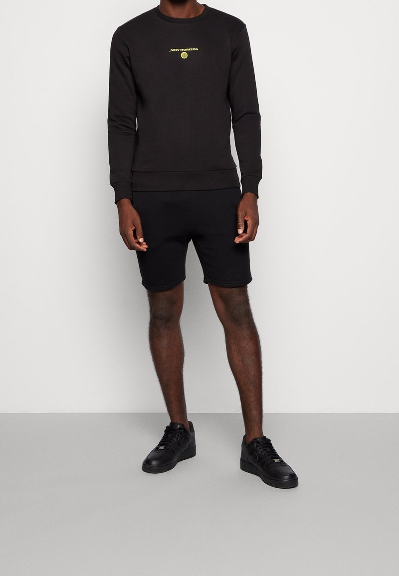 Svart sweatshirt med rund hals och långa ärmar, prydd med en gul "New Horizon"-logotyp. Matchas med svarta shorts och svarta sneakers.