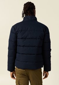 Marinblå puffjacka med hög krage, horisontellt kviltad design och elastiska ärmslut, parat med olivgröna cargobyxor.