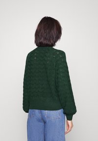 Grüner Strickpullover mit strukturiertem Muster, gerippten Bündchen und Saum. Gesehen von hinten, getragen mit hellblauen, hoch taillierten Jeans.