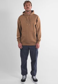 Sweat à capuche marron avec poche avant et cordon de serrage, assorti à un pantalon cargo bleu marine et des baskets noires avec des accents blancs.