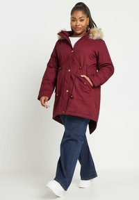 Burgundy parka takki, jossa on tekoturkishuppu, kiristettävä vyötärö, kaksi sivutaskua ja kultaiset yksityiskohdat. Päälläsin sinisiä farkkuja ja valkoisia kenkiä.