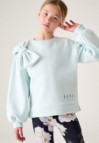 Lichtblauwe sweatshirt met een grote strik op de linker schouder, geribbelde boorden en een metallic logo op de zoom. Gecombineerd met bloemenleggings.