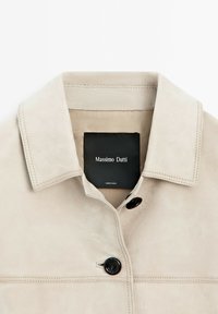 Beige mockaskinnjacka med krage, som har en svart knäppning och synliga sömmar. Inkluderar en svart etikett med "Massimo Dutti."