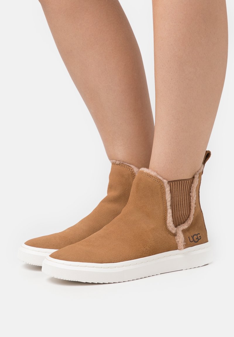 Jambes portant des bottines en daim beige avec bordure en peau lainée et semelles en caoutchouc blanches sur un fond clair uni.