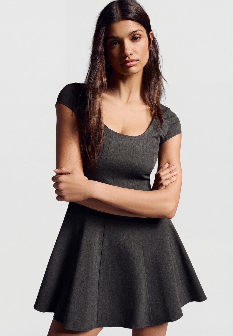Jeune femme aux longs cheveux bruns portant une robe évasée à manches courtes de couleur gris foncé, debout les bras croisés devant un fond uni.