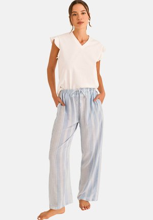 Femme portant un haut blanc à manches courtes avec col en V et un pantalon ample à rayures bleues claires avec cordon de serrage, debout pieds nus, les mains dans les poches.