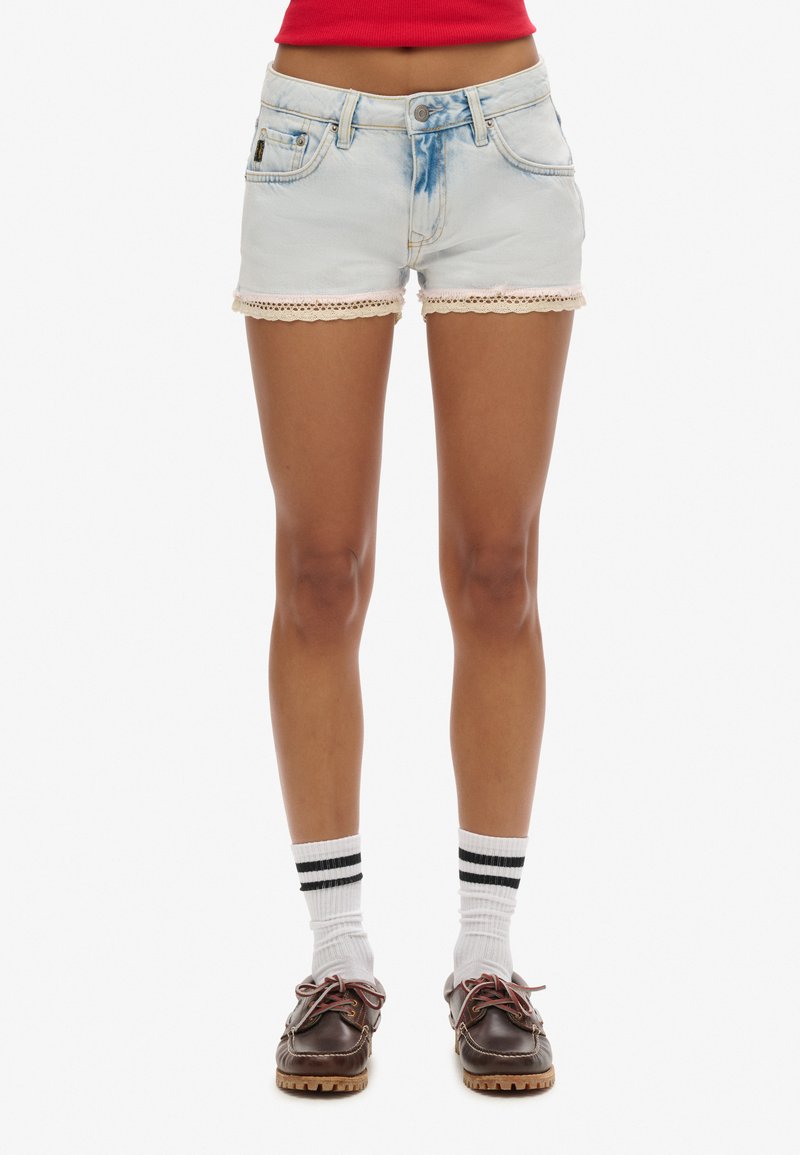 Shorts de mezclilla en azul claro con dobladillo deshilachado y trim de encaje. Cuenta con bolsillos frontales y cierre de botón. Se llevan con calcetas blancas y zapatos marrones.