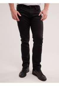 Férfi szűk fekete farmert és fekete fűzős cipőt visel, egyszerű világos háttér előtt áll.