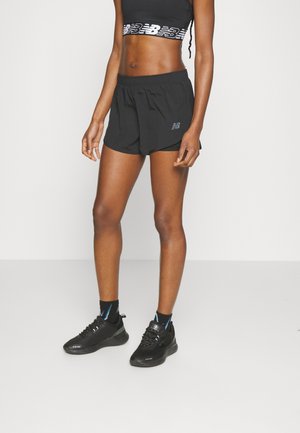 Femme portant un short de sport noir, un soutien-gorge de sport noir avec logo, des chaussettes noires et des chaussures de course noires, debout sur un sol blanc.