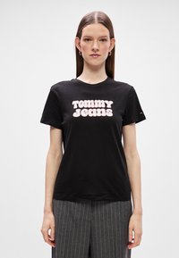 Μαύρο βαμβακερό μπλουζάκι με μεγάλη ροζ γρα�φική παράσταση "TOMMY JEANS", στρογγυλή λαιμόκοψη και κοντά μανίκια, συνδυασμένο με γκρι ρίγες παντελόνι.