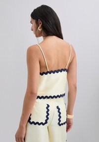Top corto amarillo con canalé y ribete en azul marino con forma de ondas, con tirantes finos tipo espagueti. Combinado con pantalones de tiro alto a juego. Vista trasera.