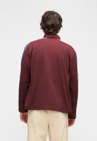 Bordowa kurtka z wysokim kołnierzem, kontrastowymi niebieskimi panelami po bokach i subtelną fakturą. Kieszenie na suwak przy dolnym brzegu.