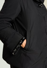 Chaqueta acolchada negra con textura suave, adornada con brillantes decoraciones negras en los puños y un cierre de botón en el cuello.