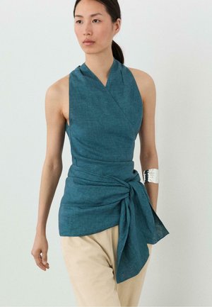 Kvinde iført ærmeløs turkis slå-om-top bundet i taljen med beige bukser og sølv manchetarmbånd på venstre arm mod en ensfarvet baggrund.