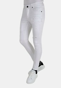 Witte skinny jeans met beschadigde details, een gladde textuur, vijf-pocket ontwerp en zwarte hardware; gecombineerd met witte sneakers.