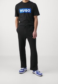 Schwarzes T-Shirt mit blauem "HUGO"-Logo, kombiniert mit schwarzen Jogginghosen mit einem Kordelzugbund und seitlichen Reißverschlüssen, ergänzt durch blaue und weiße Sneakers.